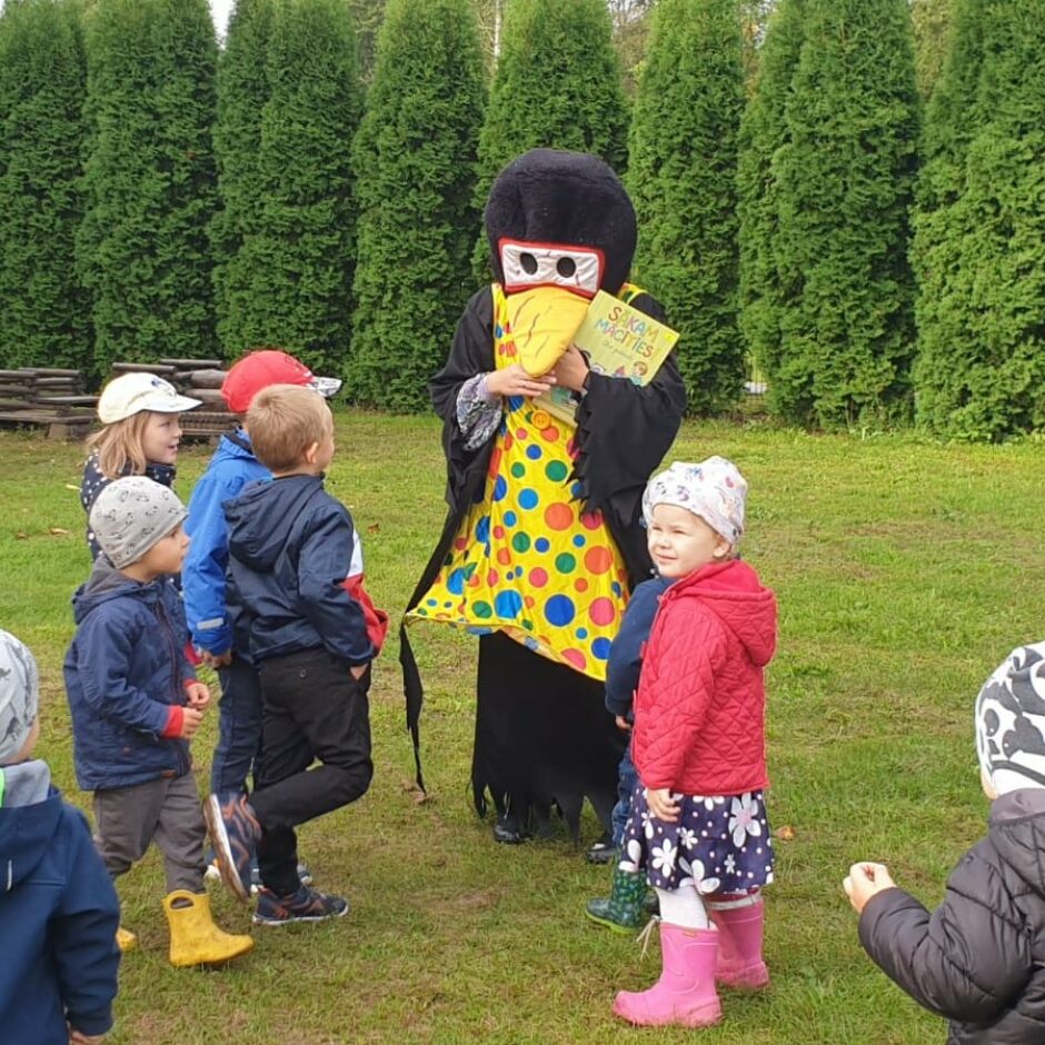 Pirmsskolas bērni āra laukumā kopā ar grupas skolotāju un maskā tērptu 