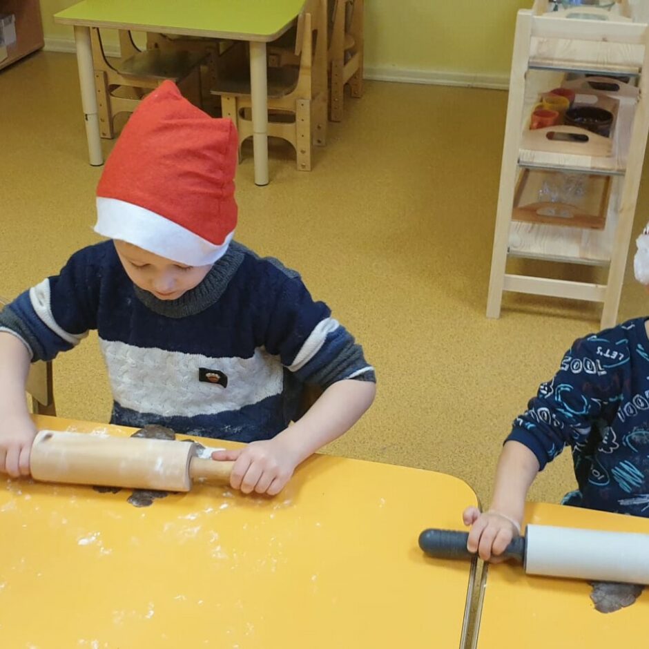 Bērni sēž pie galda un veltnē piparkūku mīklu.