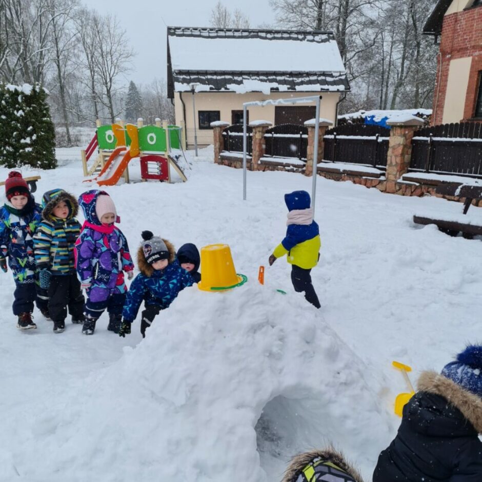 Bērni āra laukumā pie pašu veidotas sniega mājas.