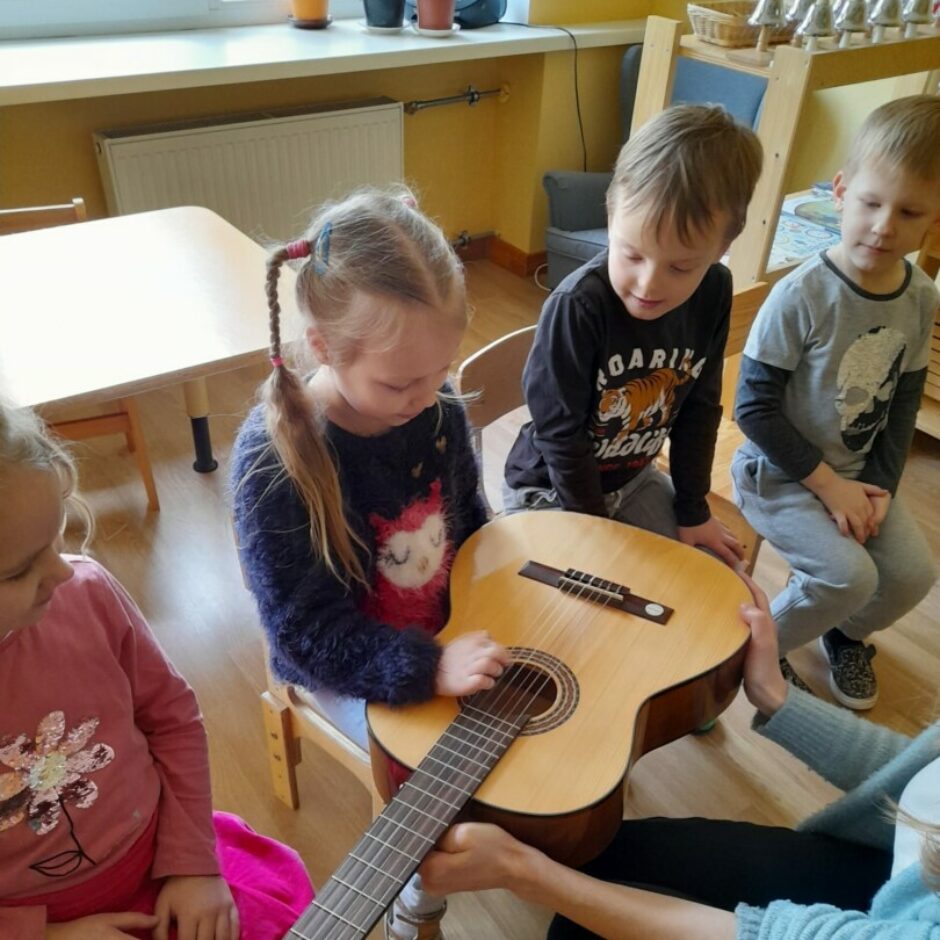 Meitene izmēģina spēlēt uz ģitāras stīgām. Citi bērni vēro.
