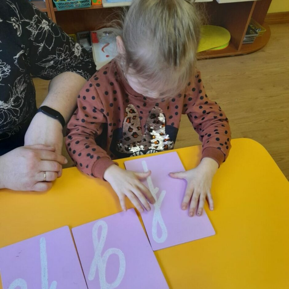 Meitene ar pirkstiem pārvelk smilšpapīra burtus.