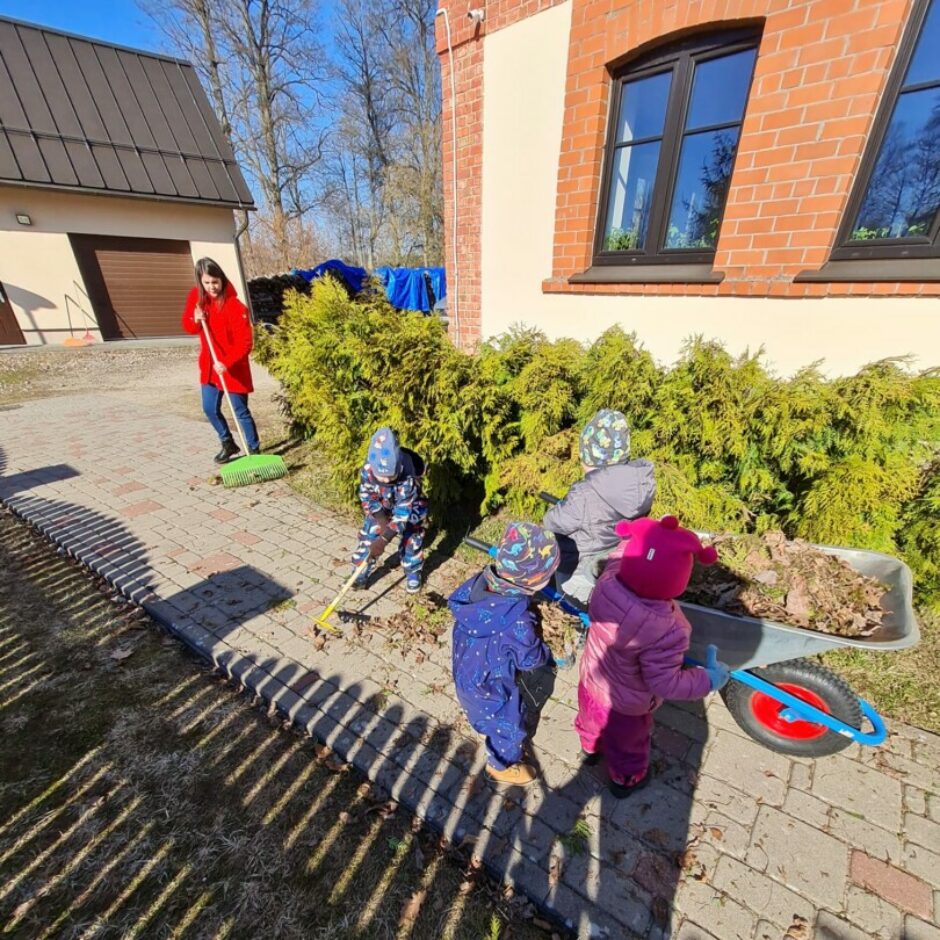 Bērni ar skolotāju sakopj apkārtni - grābj lapas un liek ķerrā.