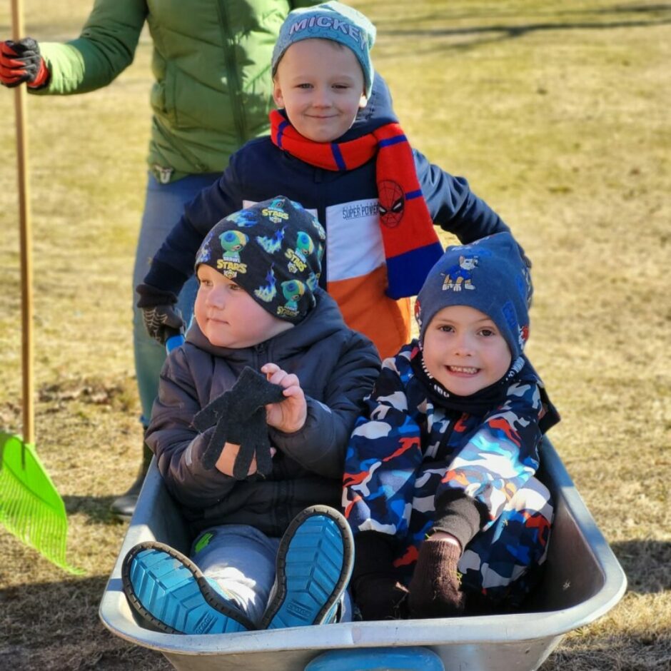 Priecīgi bērni ar ķerru - divi sēž iekšā, viens tur aiz rokturiem.