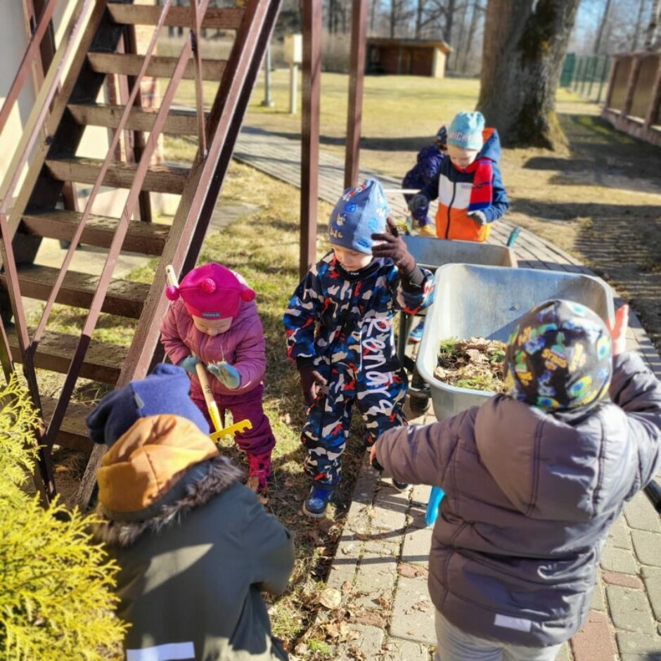 Bērni sakopj apkārtni - grābj lapas un liek ķerrā.