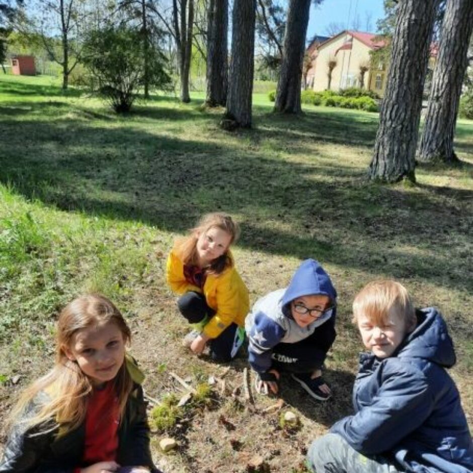 Četri skolēni veido dabas gleznu uz zemes mežā