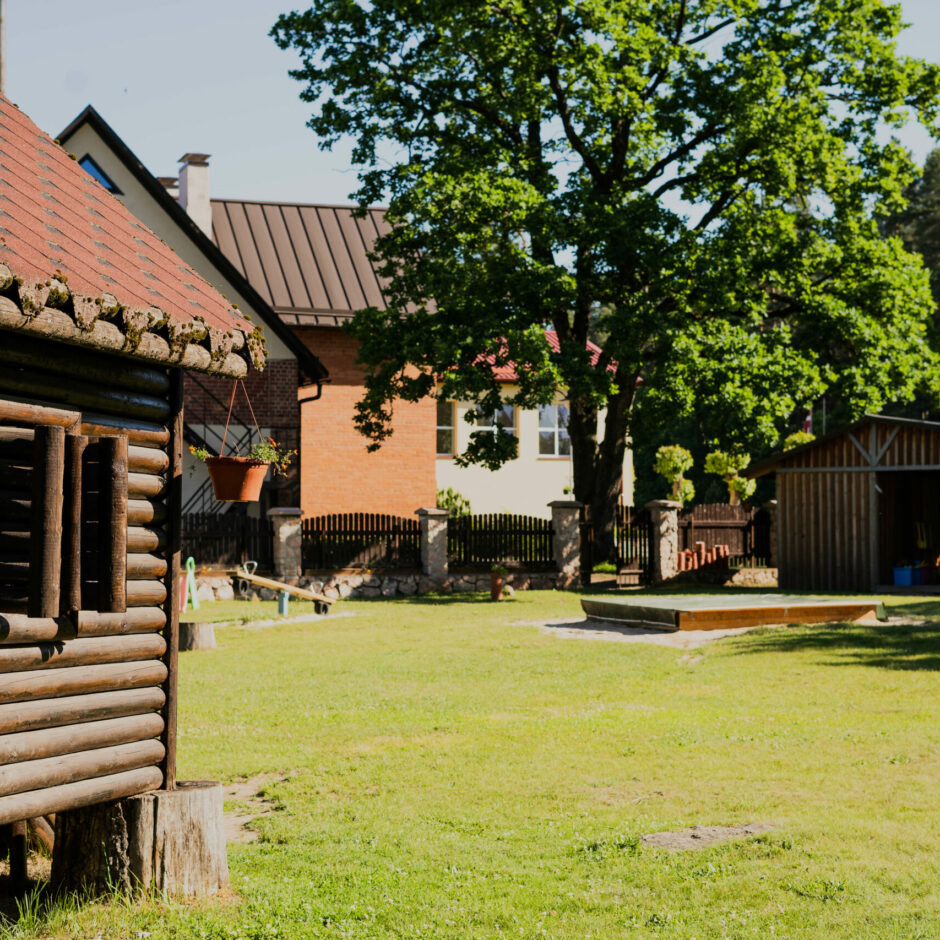 Pirmsskolas rotaļu laukums ar koka namiņu, smilškasti, nojumi un lielu ozolu laukuma malā.