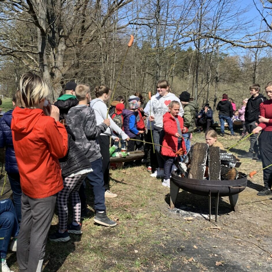 Skolēni, skolotāji stāv pie ugunskura un cep desiņas