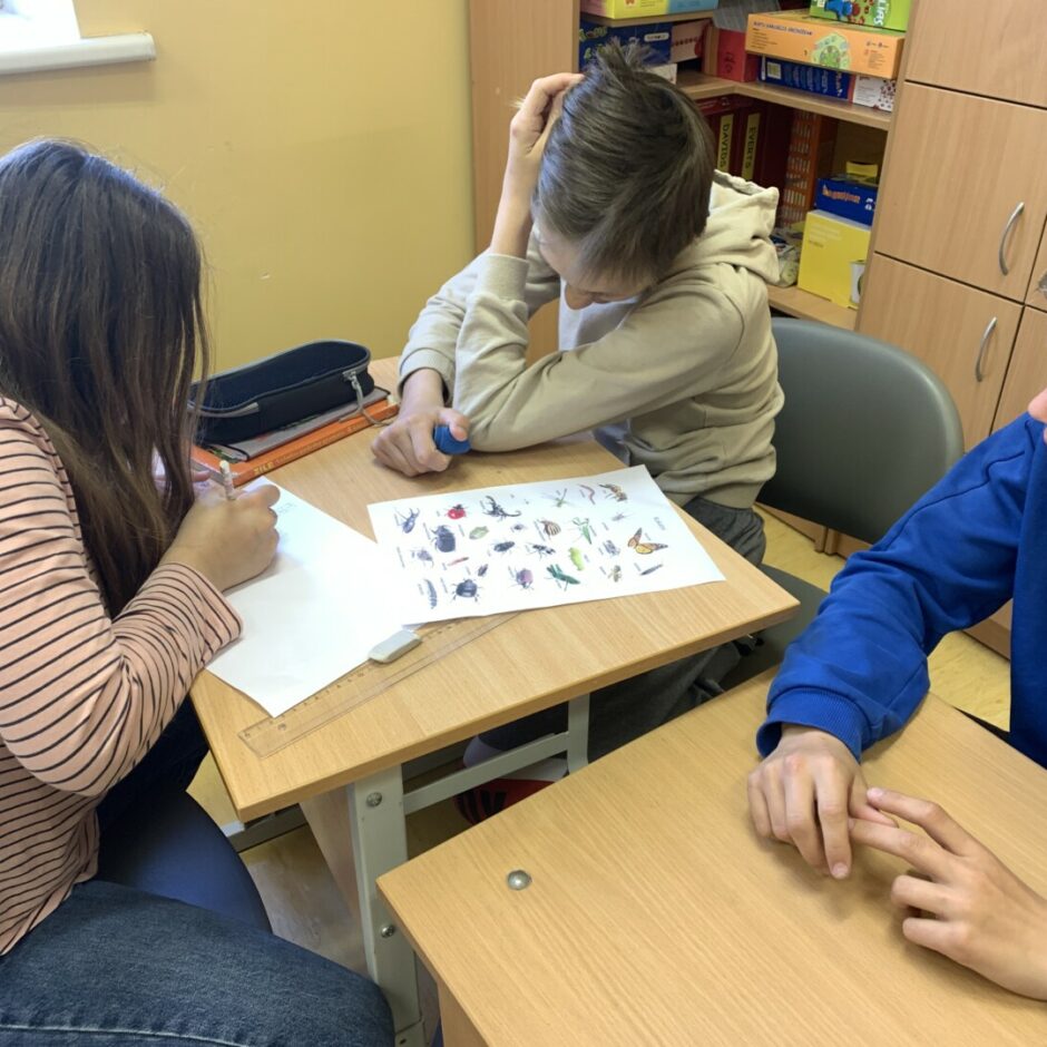 trīs bērni sēž klasē pie sola un veido mīklu