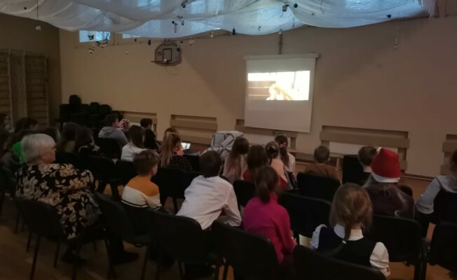 Bērni un skolotāji sēž zālē un skatās filmu