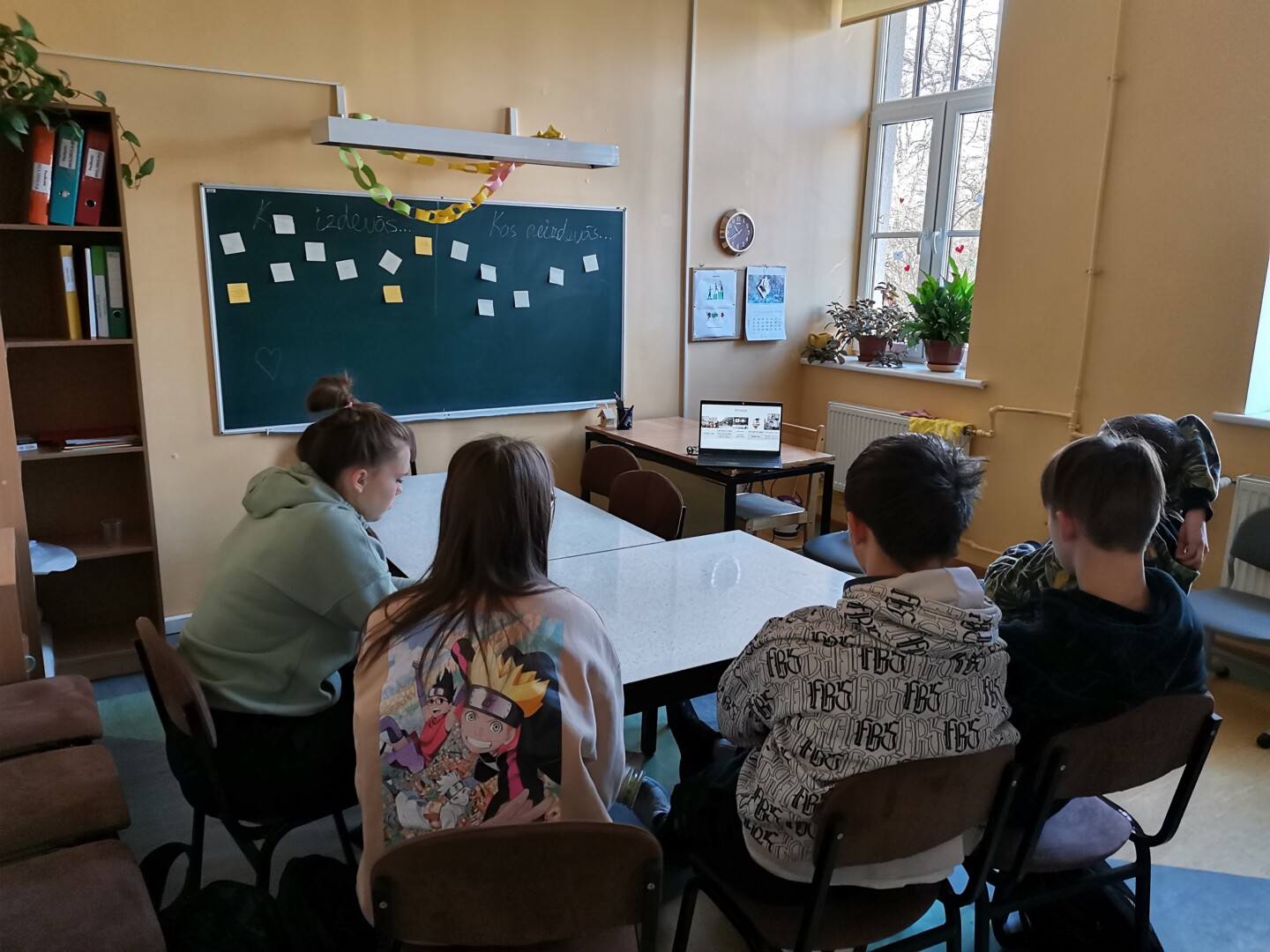 Skolēni sēž pie galda un skatās uz datoru